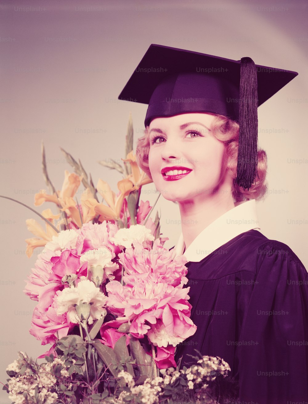 UNITED STATES - CIRCA 1950s:  Young woman wearing graduation robes and mortarboard, holding bouquet of flowers.