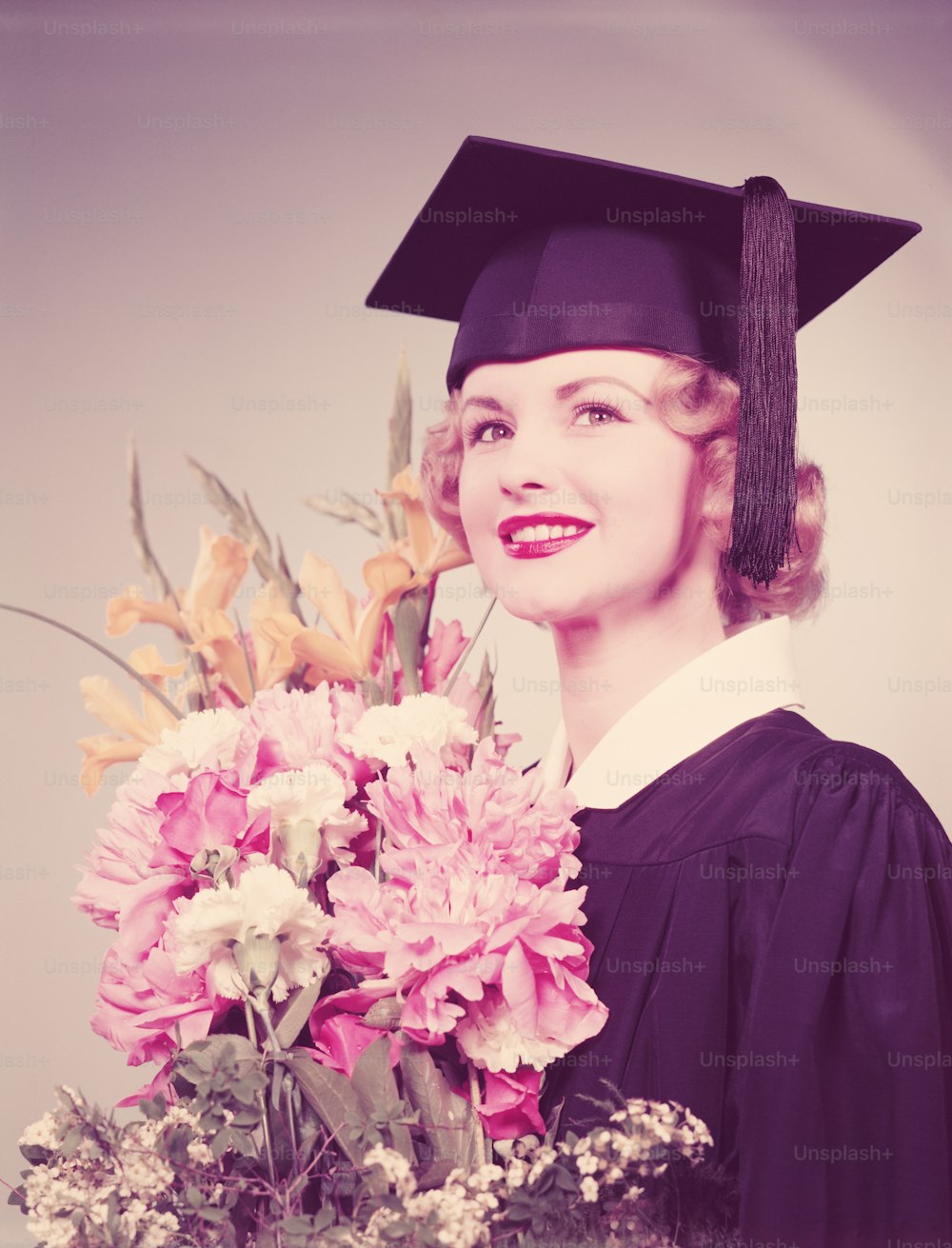 미국 - 1950년대경: 졸업 예복과 박격포를 입고 꽃다발을 들고 있는 젊은 여성.