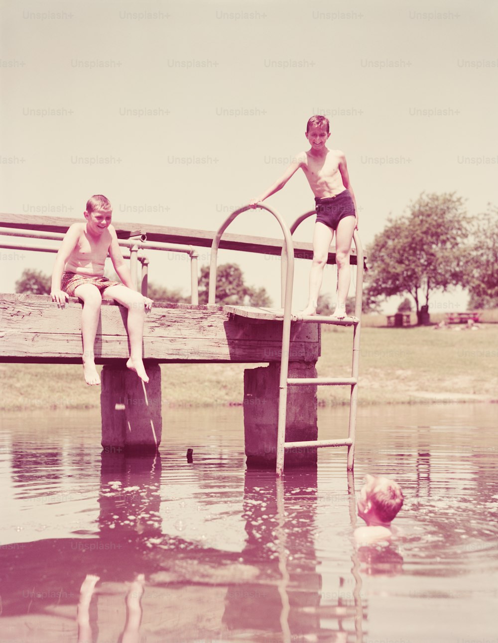 UNITED STATES - CIRCA 1950s:  Three boys at old swimming pond hole, one boy in water, one sitting on dock, one standing on ladder.