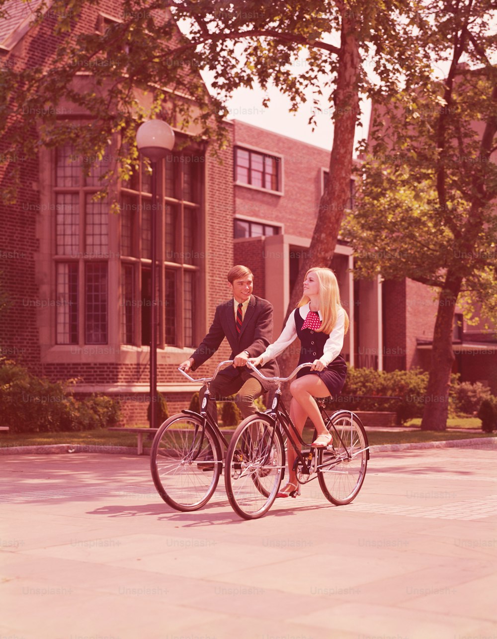 UNITED STATES - CIRCA 1960s:  Teenage couple riding bicycles past high school buildings.