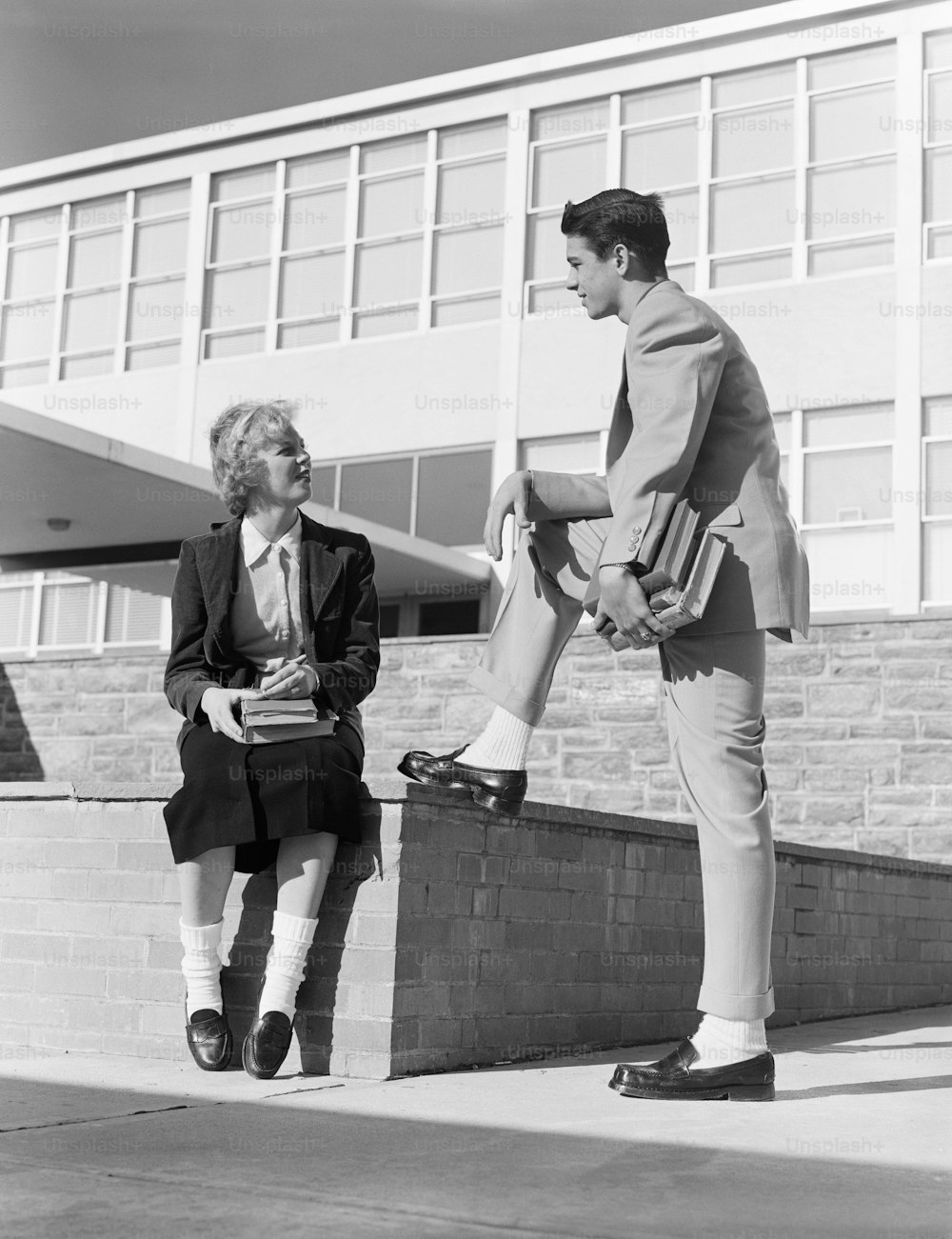 UNITED STATES - CIRCA 1950s:  High school girl sitting on wall, talking to teenage boy.
