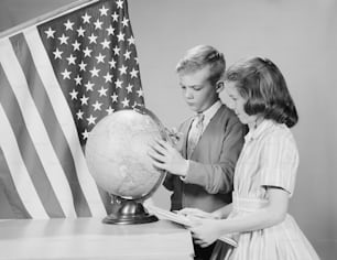 UNITED STATES - CIRCA 1960s:  Boy and girl standing at desk, looking at globe, American flag in background.