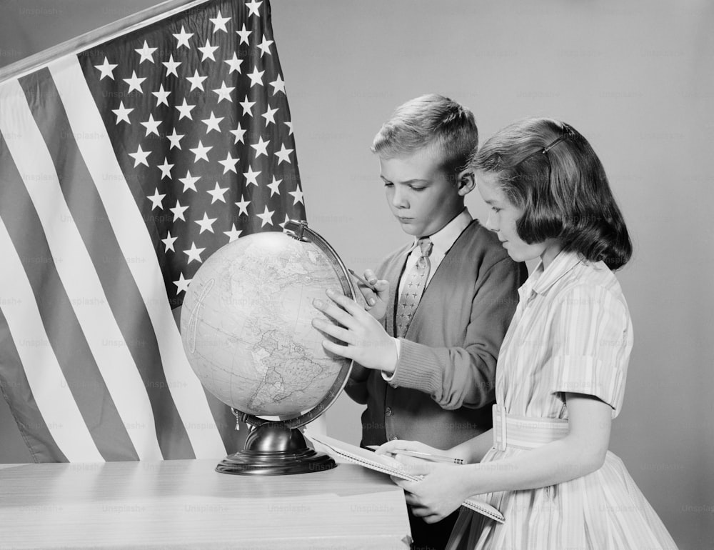 UNITED STATES - CIRCA 1960s:  Boy and girl standing at desk, looking at globe, American flag in background.
