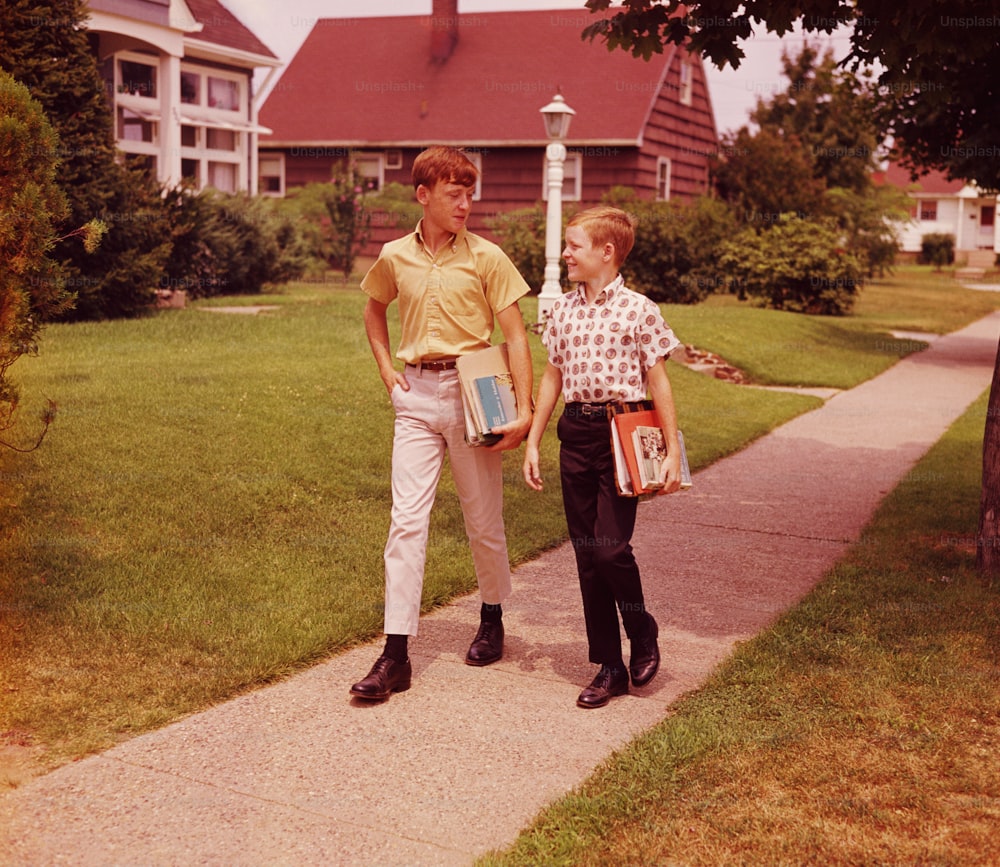 UNITED STATES - CIRCA 1960s:  Two boys walking along street, talking, carrying school books.
