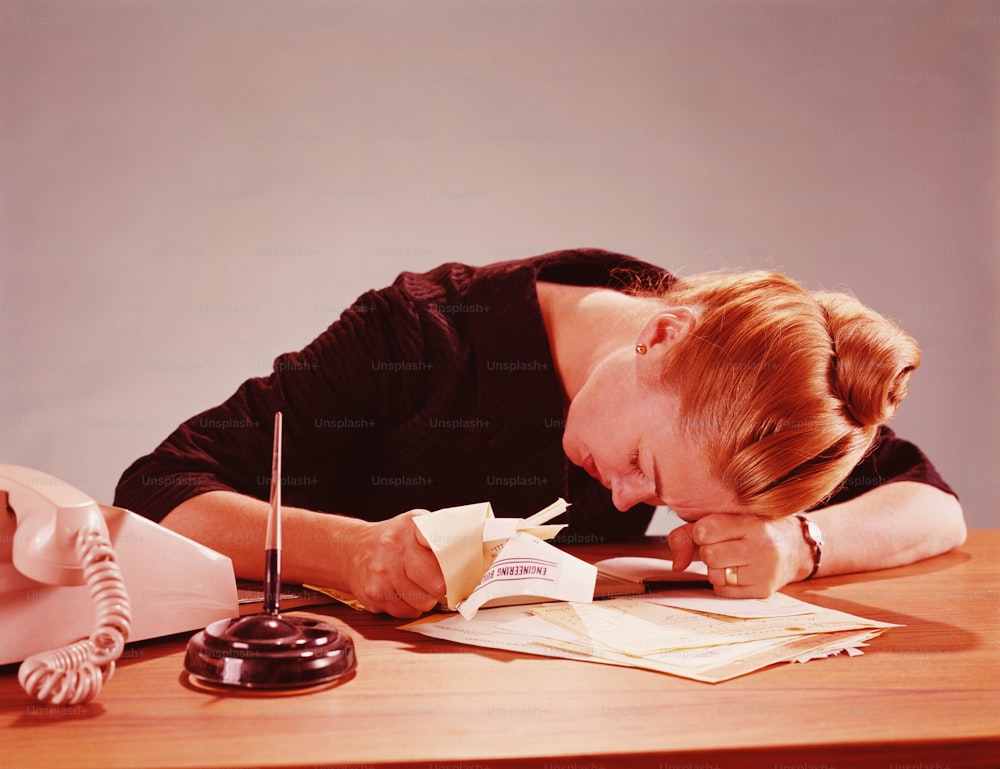 UNITED STATES - CIRCA 1960s:  Unhappy young woman slumped across desk over papers, grasping some in hand.