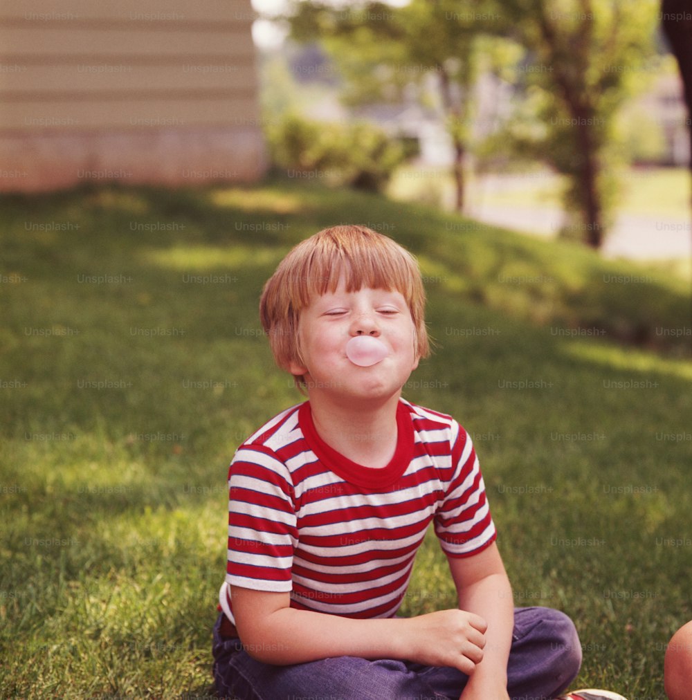 미국 - 1960년대경: 풀밭에 앉아 풍선껌 거품을 불고 있는 소년.