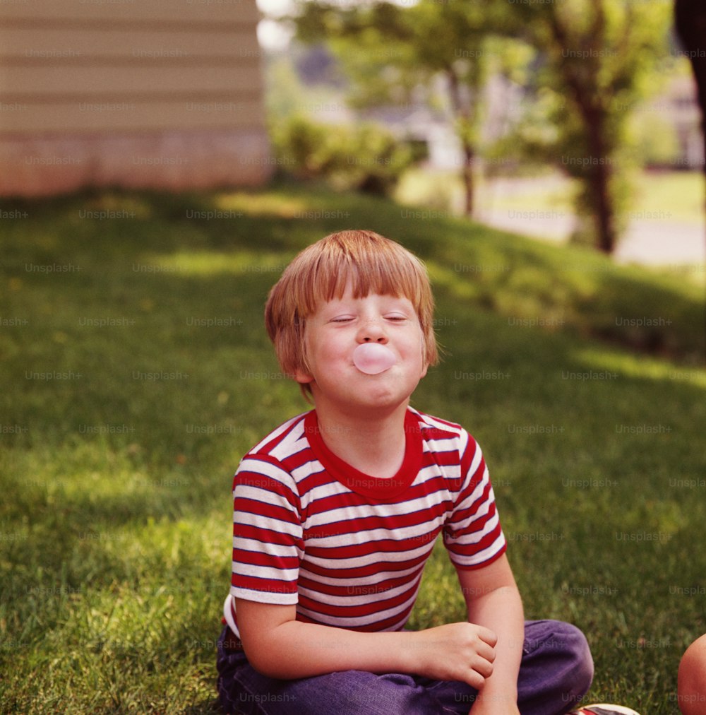 UNITED STATES - CIRCA 1960s:  Boy sitting in grass, blowing bubblegum bubble.