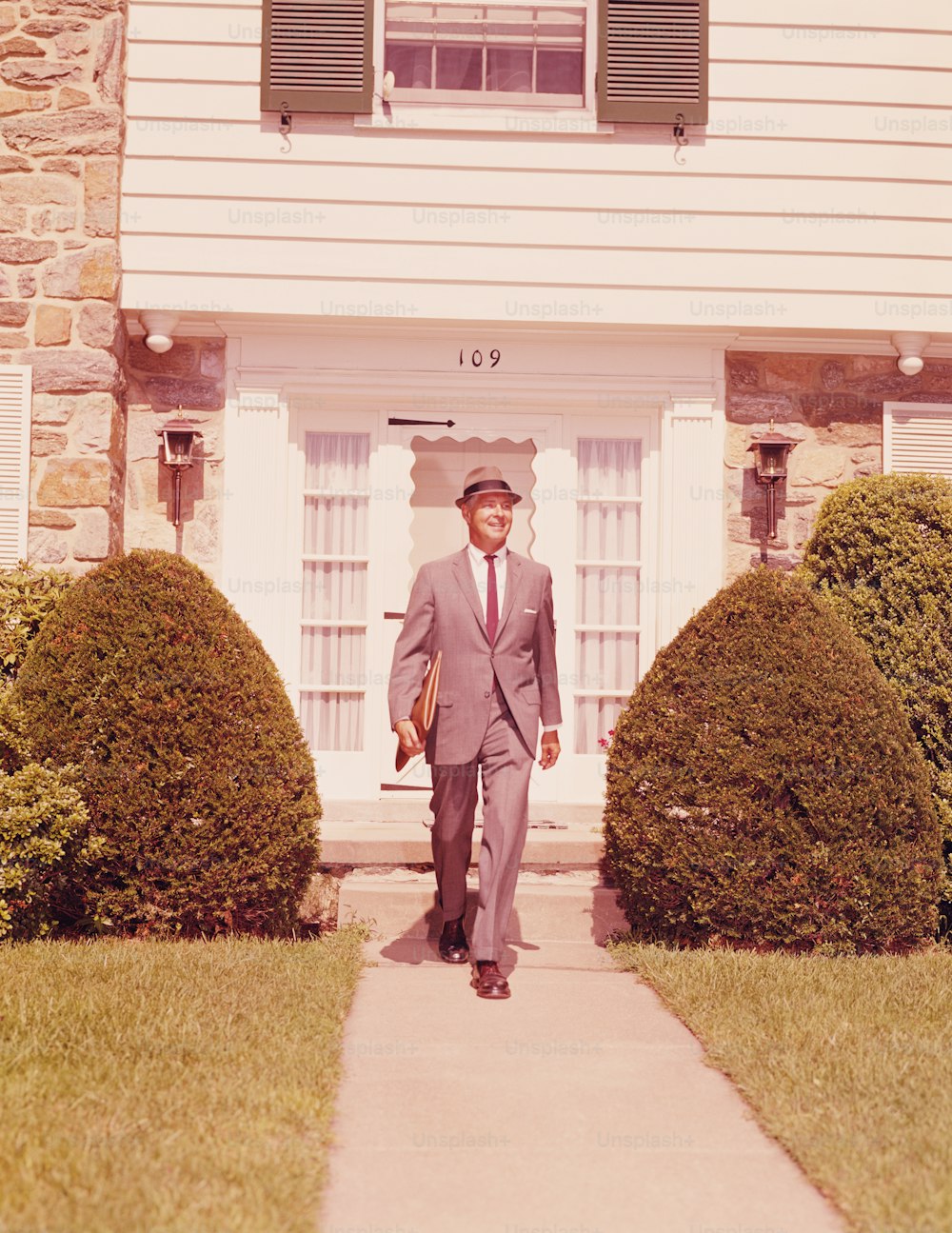 UNITED STATES - CIRCA 1960s:  Salesman wearing hat and carrying briefcase, walking along suburban street.