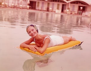 ESTADOS UNIDOS - POR VOLTA DE 1970: Mulher na piscina reclinada em jangada inflável, usando touca de banho, sorrindo.