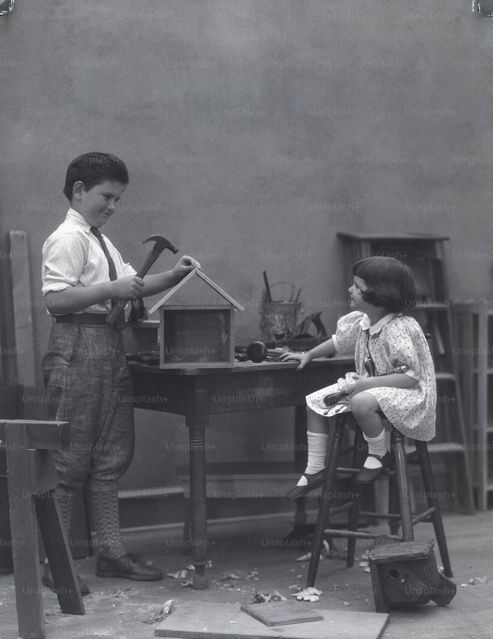 UNITED STATES - CIRCA 1930s:  Boy and girl in workshop, girl watching as boy hammers nail into birdhouse.