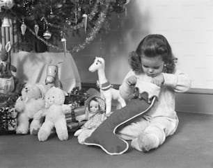 VEREINIGTE STAATEN - CIRCA 1940er Jahre: Junges Mädchen im Schlafanzug, das einen Weihnachtsstrumpf neben dem Weihnachtsbaum öffnet.