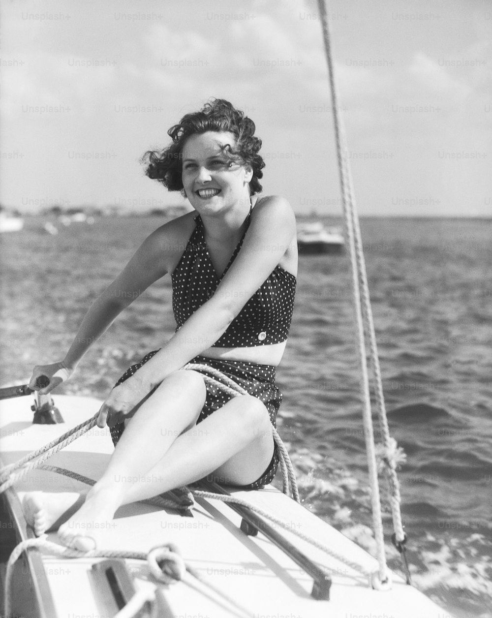 UNITED STATES - CIRCA 1930s:  Woman in swimsuit, on sailing boat, holding rope, smiling.