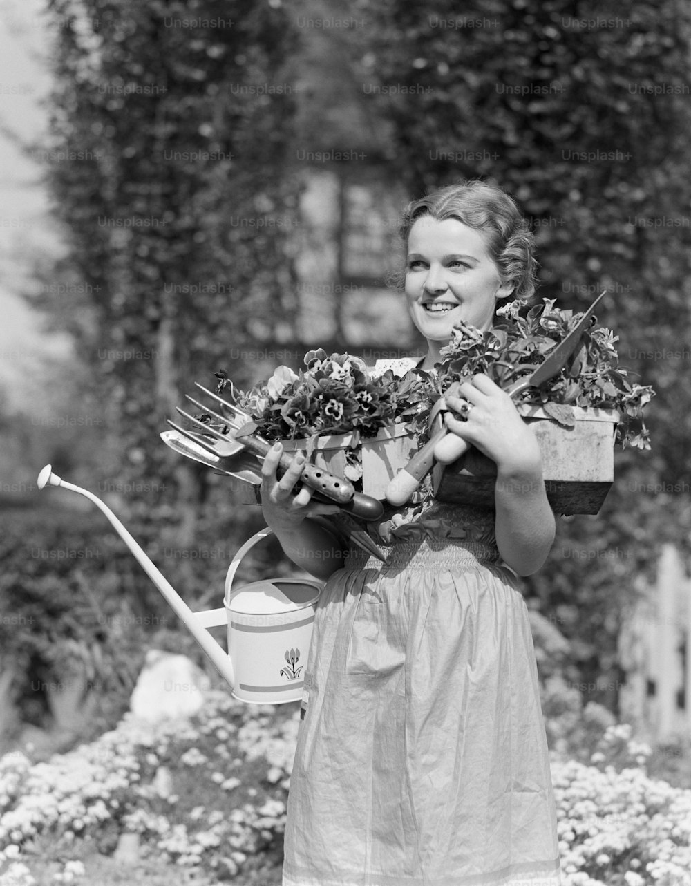 UNITED STATES - CIRCA 1930s:  Woman standing in garden, smiling, arms full carrying watering can, garden tools and potted plants, portrait.