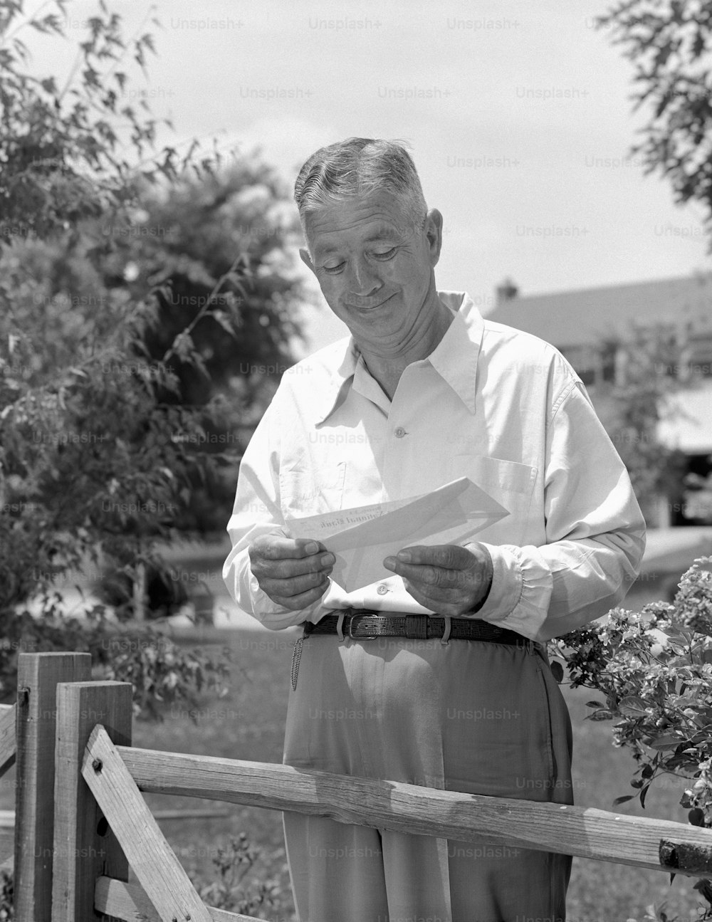 UNITED STATES - CIRCA 1950s:  Elderly man standing by fence in yard, opening envelope and taking out cheque.
