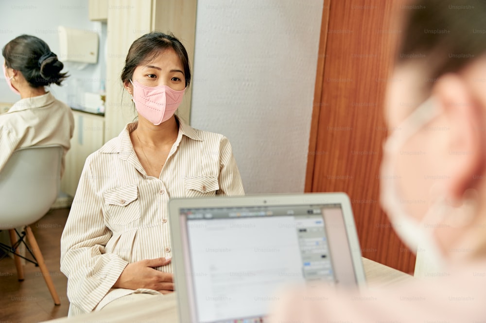 a woman sitting in front of a laptop computer wearing a pink face mask