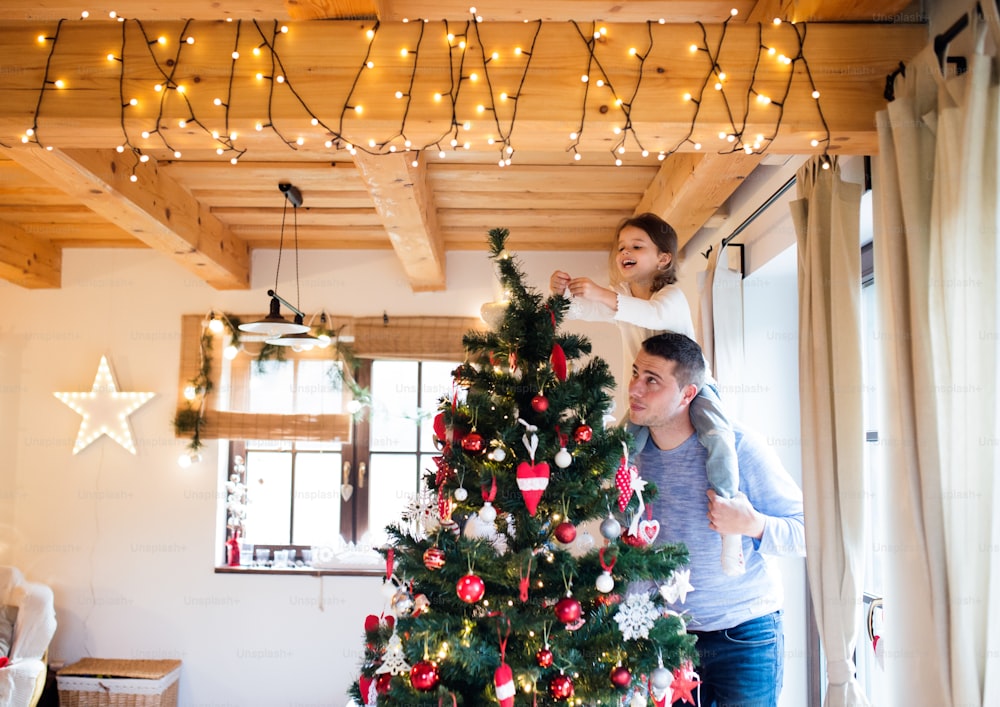 Padre joven dando a su pequeña hija a cuestas decorando el árbol de Navidad juntos.