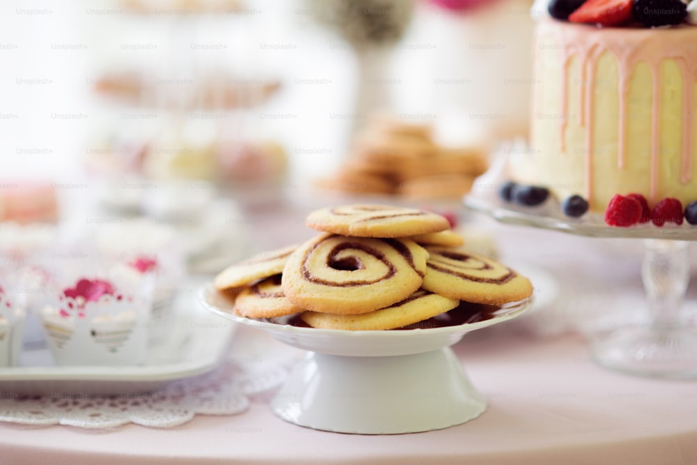 Galletas en forma de espiral, bizcocho con frutos rojos y cupcakes colocados sobre mesa con mantel rosa y encaje artesanal. Toma de estudio.