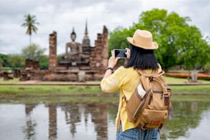 아시아 관광 여성은 모바일을 사용하여 태국 수코타이 (Sukhothai)에서 태국 건축물의 고대 탑 사원 사진을 찍습니다. 캐주얼 태국 옷감 스타일의 여성 여행자 방문 도시 개념