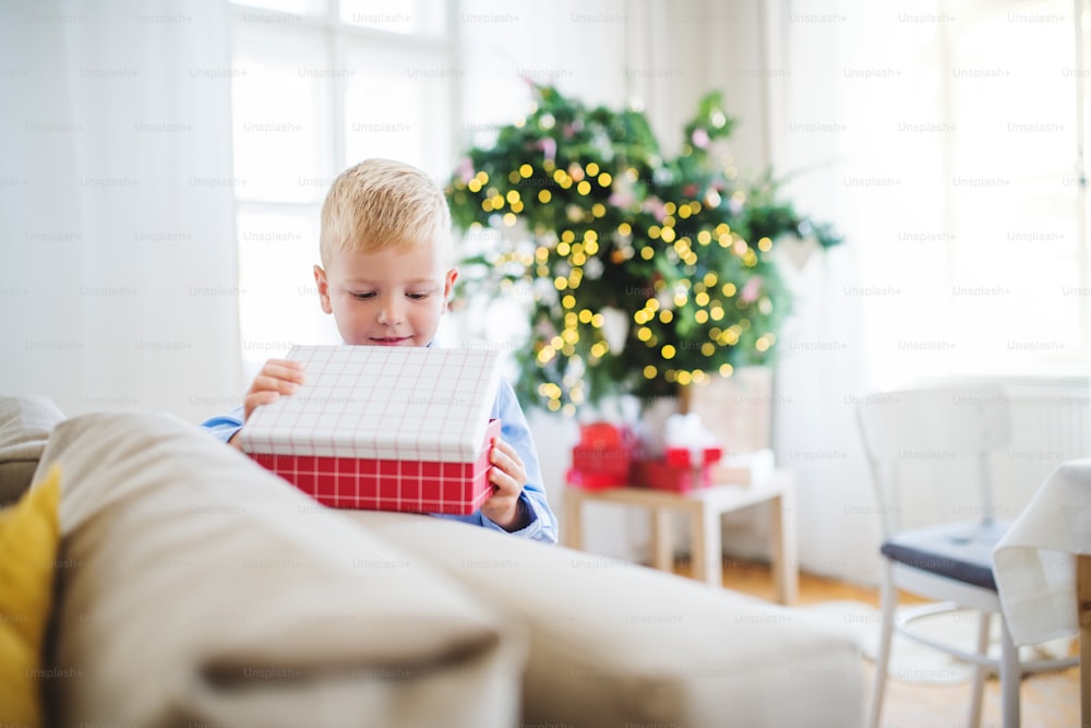Un niño pequeño de pie junto a un sofá en casa en Navidad, abriendo un regalo.
