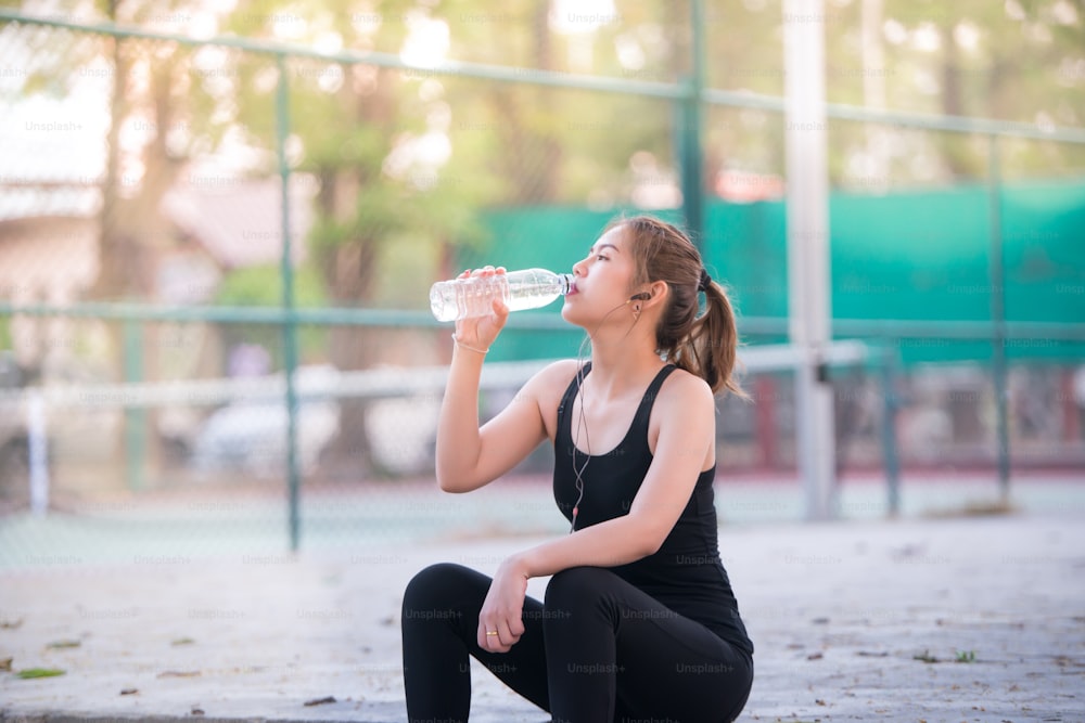 Asiatische junge Fitness-Frau trinkt Wasser nach dem Joggen mit Tennisplatzhintergrund