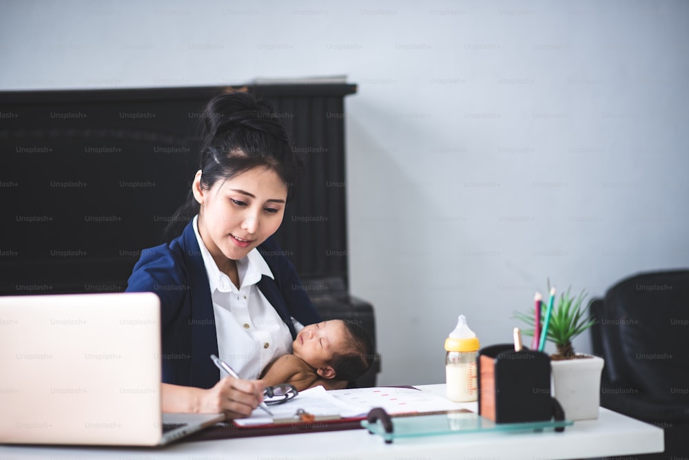 Vielbeschäftigte junge Frau, die am Laptop arbeitet oder studiert, während sie ihr Baby zu Hause in den Armen hält
