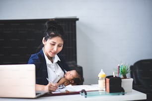Giovane donna occupata che lavora o studia sul computer portatile mentre tiene il suo bambino in braccio a casa