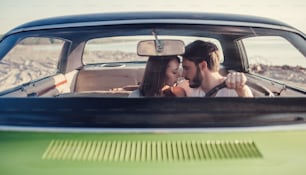 Romantic couple is sitting in green retro car on the beach. Handsome bearded man and attractive young woman in vintage classic car. Stylish love story. Hugging and kissing while being in car.