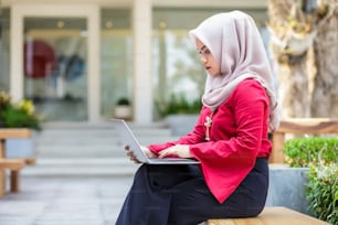 Junge asiatische muslimische Frau, die im Freien am Laptop arbeitet.