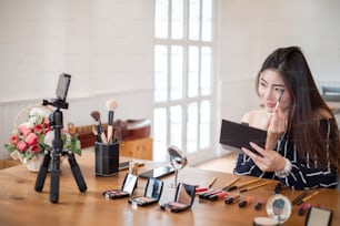 Bloguera asiática con cosméticos de maquillaje grabando videoclip con teléfono inteligente en casa.