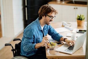 Young happy entrepreneur in wheelchair surfing the net on laptop while healthy food at home.