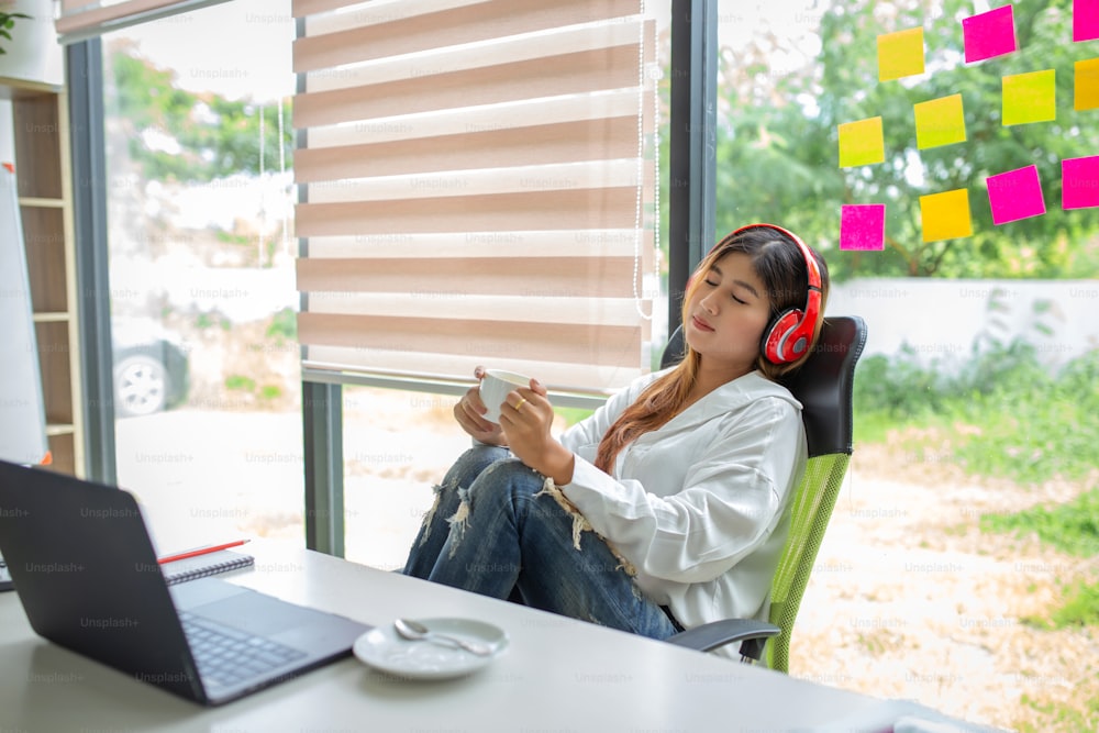 Portrait of young asian woman listening to music with headphones and using laptop computer and drinking coffee at office.