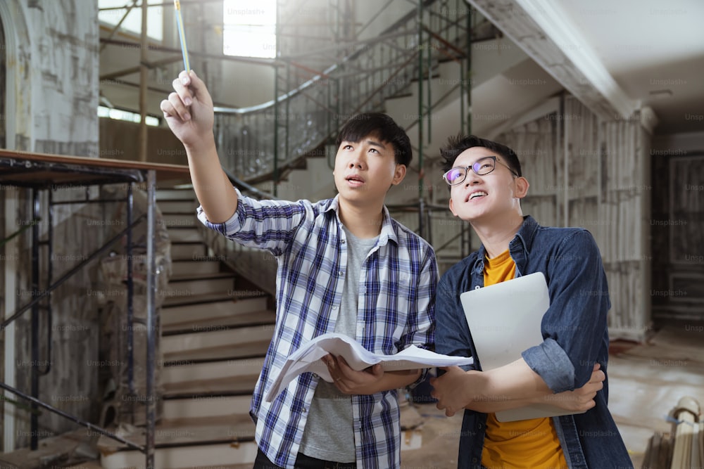 Dos contratistas masculinos de interiores asiáticos consultan y examinan en la renovación de la casa del sitio con computadora portátil y dibujo en papel, el ingeniero y el diseñador asiáticos explican y discuten en la construcción del sitio