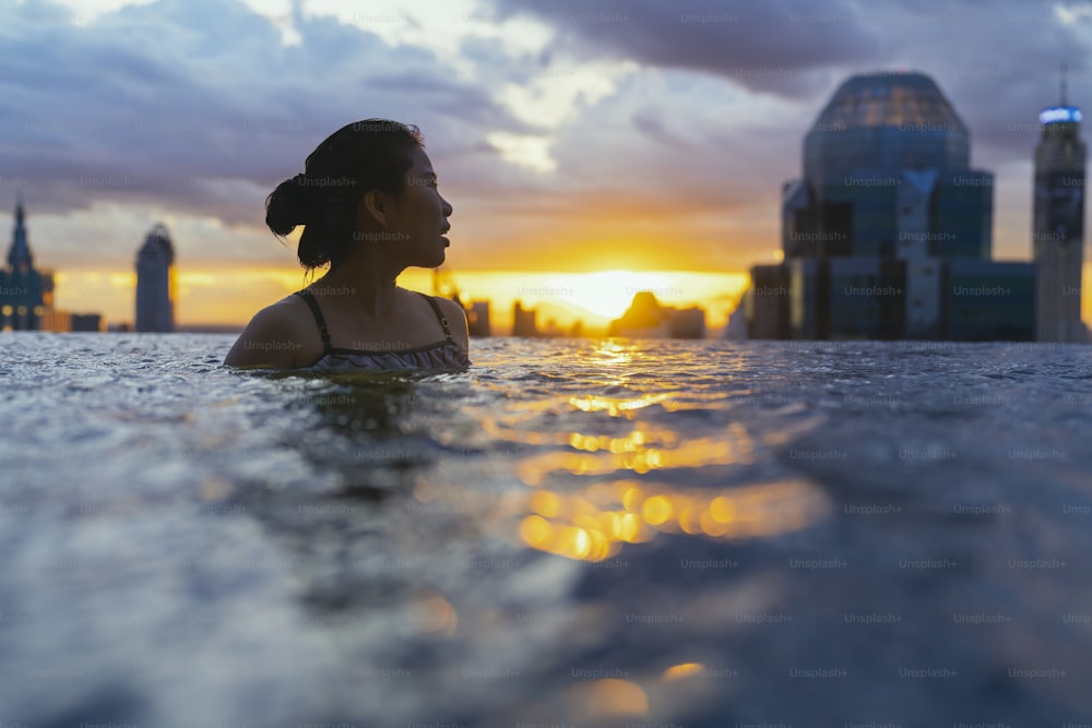 아시아 여성의 검은 실루엣은 여름 휴가 휴가에 물을 튀기고 푸른 바다 일몰 전망이 있는 인피니티 수영장에서 고층 스카이 스케이프 도시 도심 건강한 행복 라이프스타일