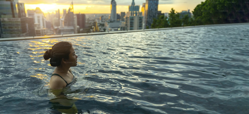 Schwarze Silhouette der asiatischen Frau spritzt Wasser im Sommerurlaub Entspannung im Infinity-Pool mit blauem Meer Sonnenuntergang mit Hochhaus Skyscape Urban Downtown Gesundes Glück Lebensstil
