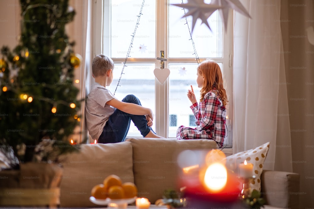 Small girl and boy in pajamas indoors at home at Christmas, sitting on window sill and talking.