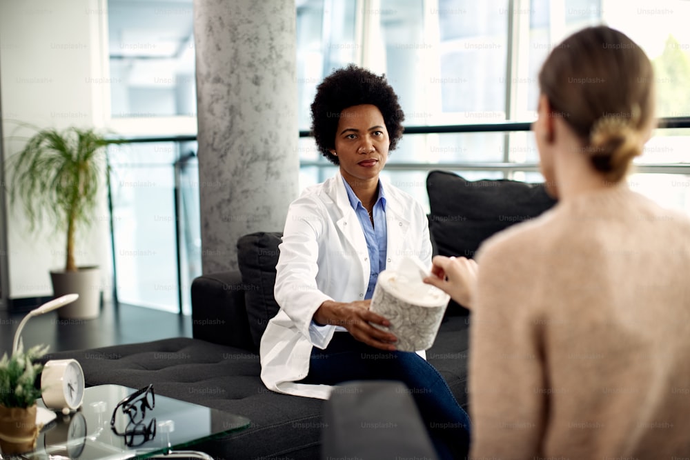 Psicoterapeuta negra dando pañuelos de papel a un paciente durante una cita en su consultorio.