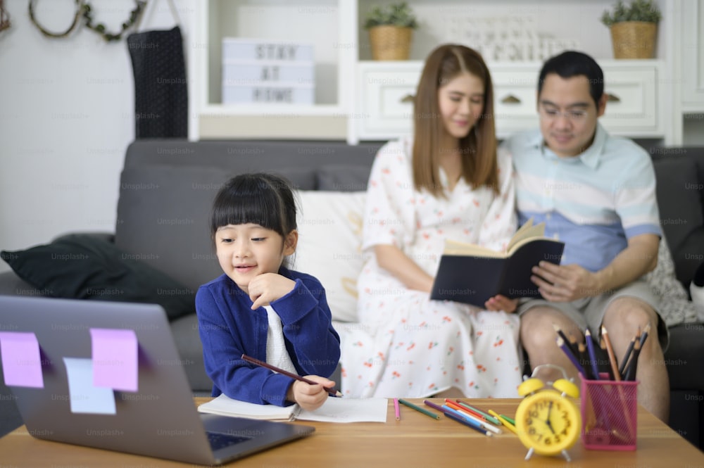 アジアの幸せな娘は、親が自宅のソファに座っている間、インターネットを介してオンラインで勉強するためにラップトップを使用しています。eラーニングのコンセプト