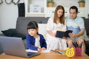 La hija feliz asiática está usando una computadora portátil para estudiar en línea a través de Internet mientras los padres están sentados en el sofá de casa. Concepto de e-learning