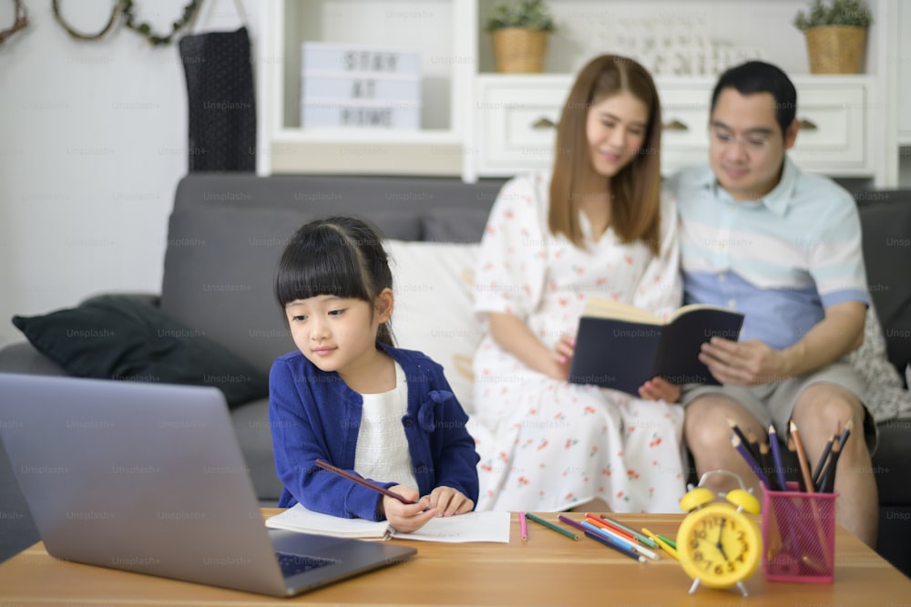 アジアの幸せな娘は、親が自宅のソファに座っている間、インターネットを介してオンラインで勉強するためにラップトップを使用しています。eラーニングのコンセプト