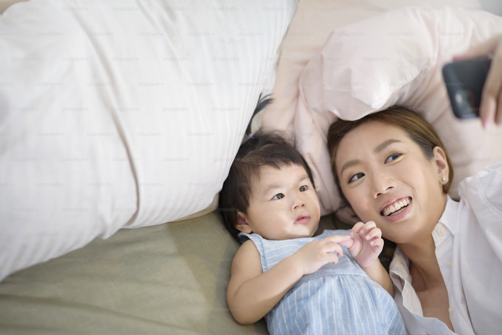 Asian Mother and her baby daughter are making selfie or video call to father in bed , Family , Home safety , parenthood , technology concept .