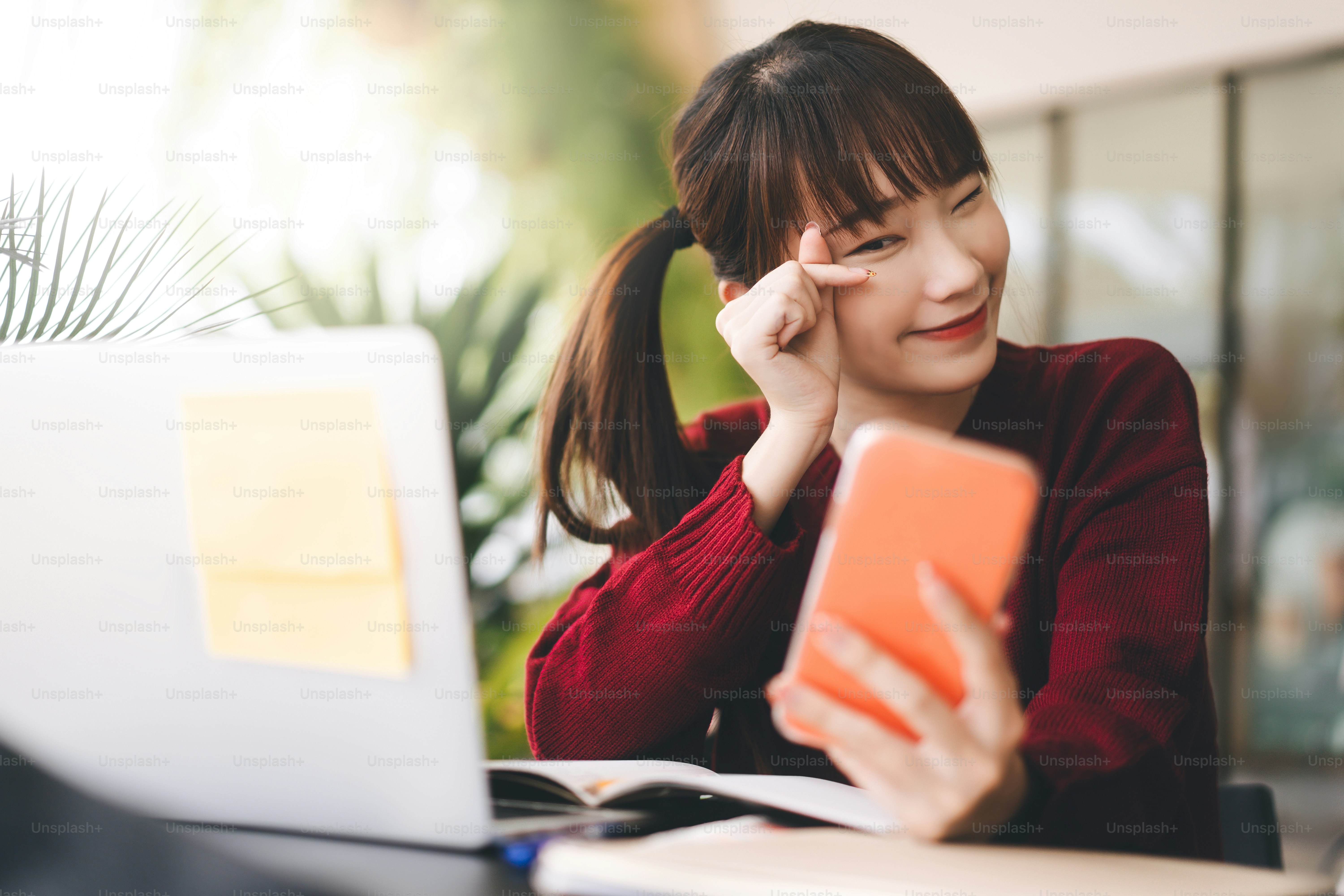 Wanita siswa asia dewasa muda yang bahagia mengenakan sweter merah. Menggunakan ponsel dan laptop untuk belajar online di kafe. Gaya hidup masyarakat universitas dengan teknologi modern pada konsep hari hari.