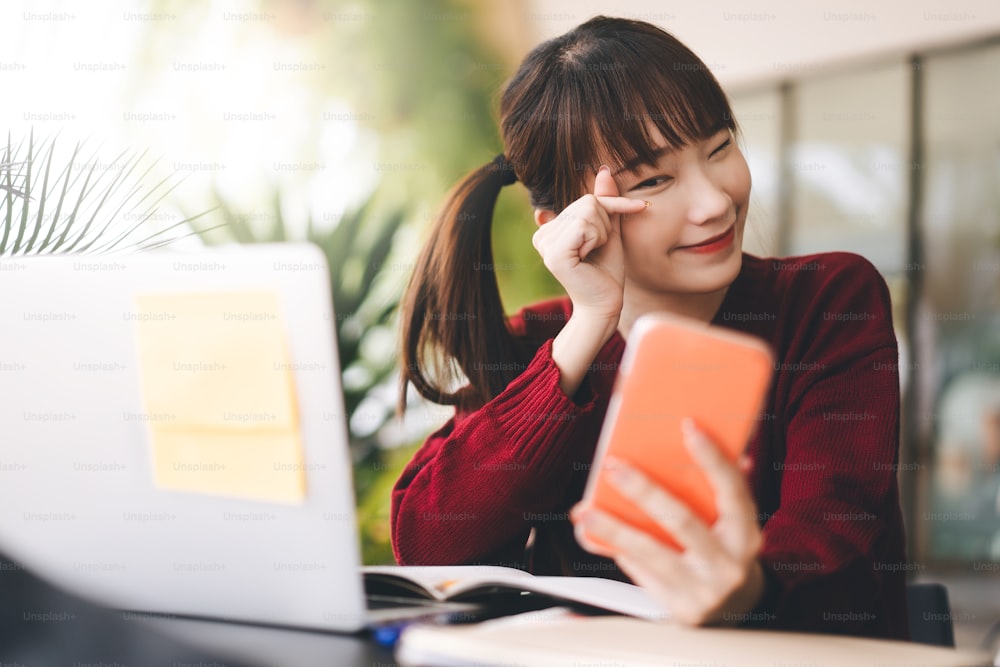 幸せな若い大人のアジアの学生女性は赤いセーターを着ています。カフェで携帯電話とノートパソコンを使ってオンラインで勉強する。現代のテクノロジーを備えた大学時代のライフスタイルをコンセプトに。