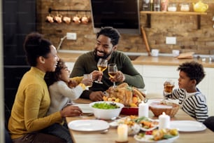 Happy black parents and kids toasting while celebrating Thanksgiving day during family lunch at dining table.