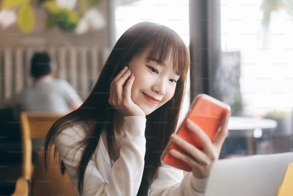 La gente universitaria, la educación, el estilo de vida, el trabajo y el estudio en el concepto diurno. Estudiante asiática adulta joven que usa el teléfono móvil para la aplicación en línea en el café interior.