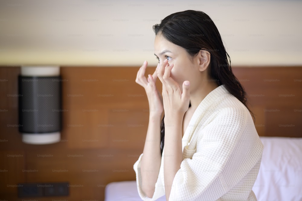 A happy beautiful woman in white bathrobe applying moisturizing cream on face in bedroom, skin care and treatment concept