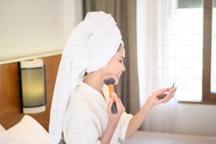 Attraente femmina felice in accappatoio bianco sta applicando il trucco naturale con il pennello della polvere cosmetica, Beauty Concept.