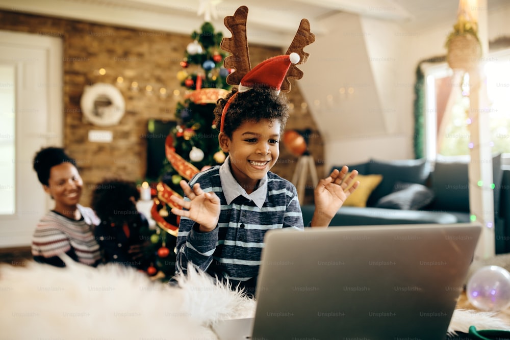 Joyful black boy using laptop and greeting someone during video call on Christmas day at home. His family is in the background.