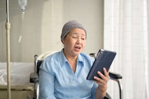 Una mujer paciente de cáncer con pañuelo en la cabeza haciendo una videollamada en la red social con familiares y amigos en el hospital.