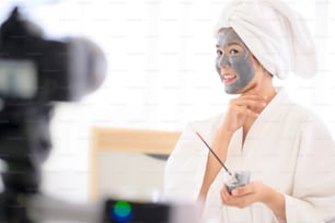 Video camera filming woman in white bathrobe applying a face mask for movie, behind the scenes of shoot