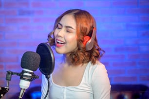 Eine junge lächelnde Sängerin mit Kopfhörern und Mikrofon, während sie in einem Musikstudio mit bunten Lichtern Songs aufnimmt.